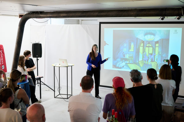 Anna Just, in kobaltblau, präsentiert ihre aktuellen Arbeiten  vor den österreichischen Verlagshäusern. im Designforum Austria. Via Beamer sieht man das Bild "Oh Tante Petunia" und dazu erzählt sie von ihrem Verständnis von Raum und Licht in ihren Bildern. Links im Bild sind auch Rosa Hirzer und Lili Richter, ebenfalls Illustratorinnen, zu sehen.